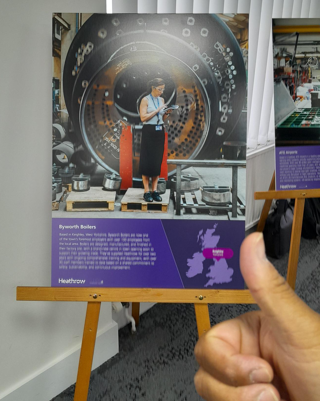 A picture easel with an image of a woman in a factory, in front on the easel is a hand with a thumbs up