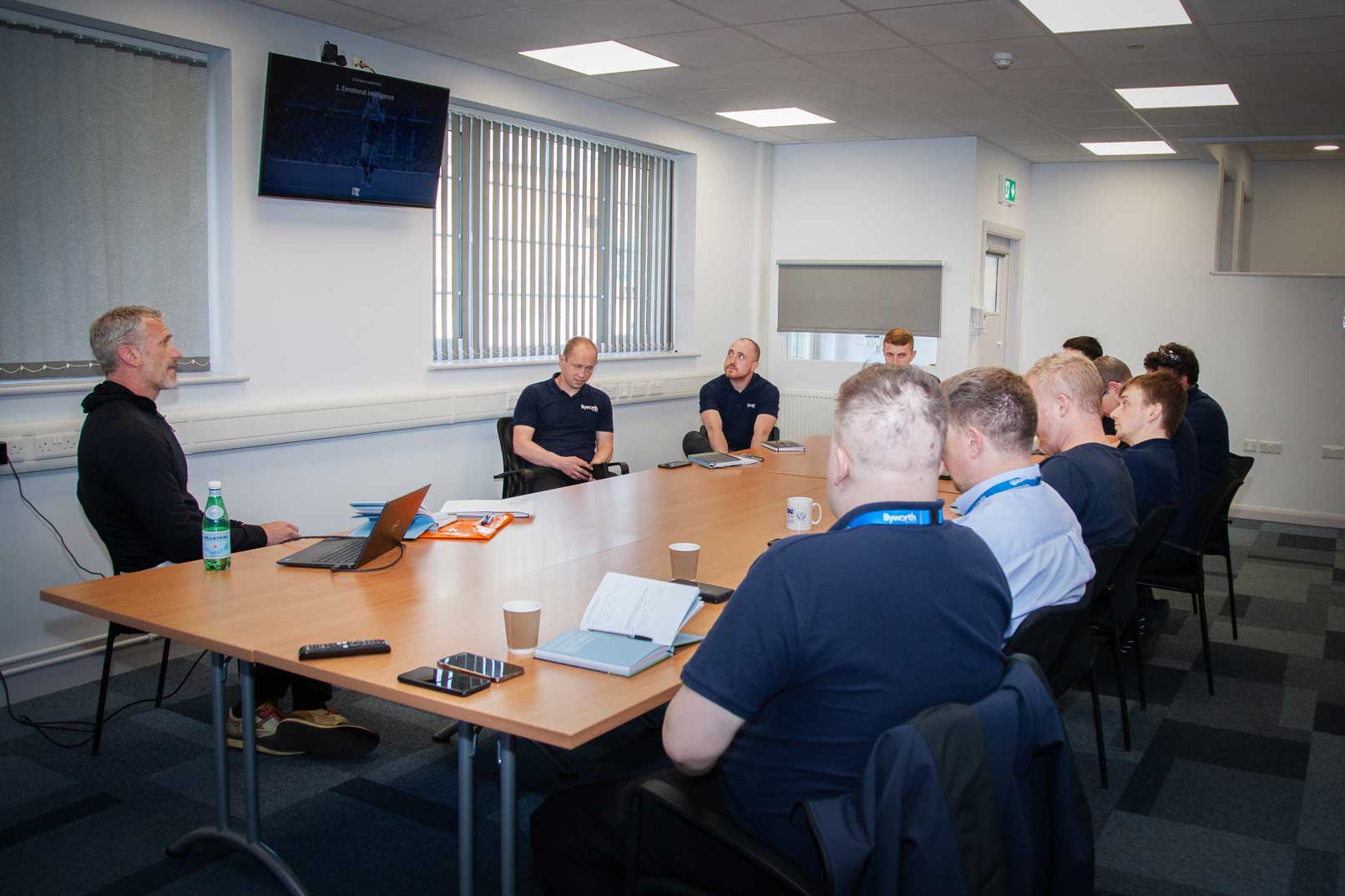 A board room with a large wooden table, men are sat around listening to each other talk