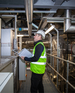 Man wearing a hard hat, high visibility vest and holding a clipboard looks up a pipework within an industrial boiler house