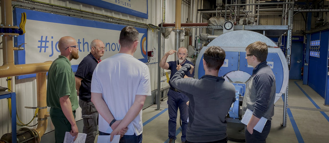 six men are stood in an industrial boiler house talking. Two men have their arms in the air showing each other something made of glass 